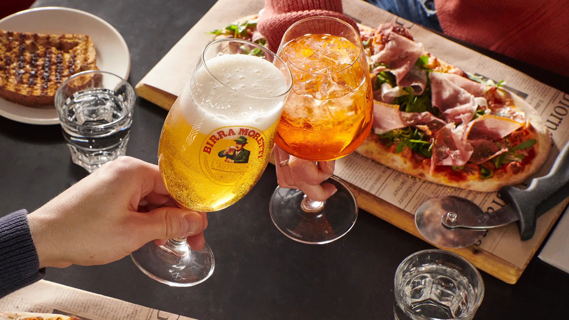 Two people toasting with beer and spritz beside a prosciutto-topped pizza at PACINI.
